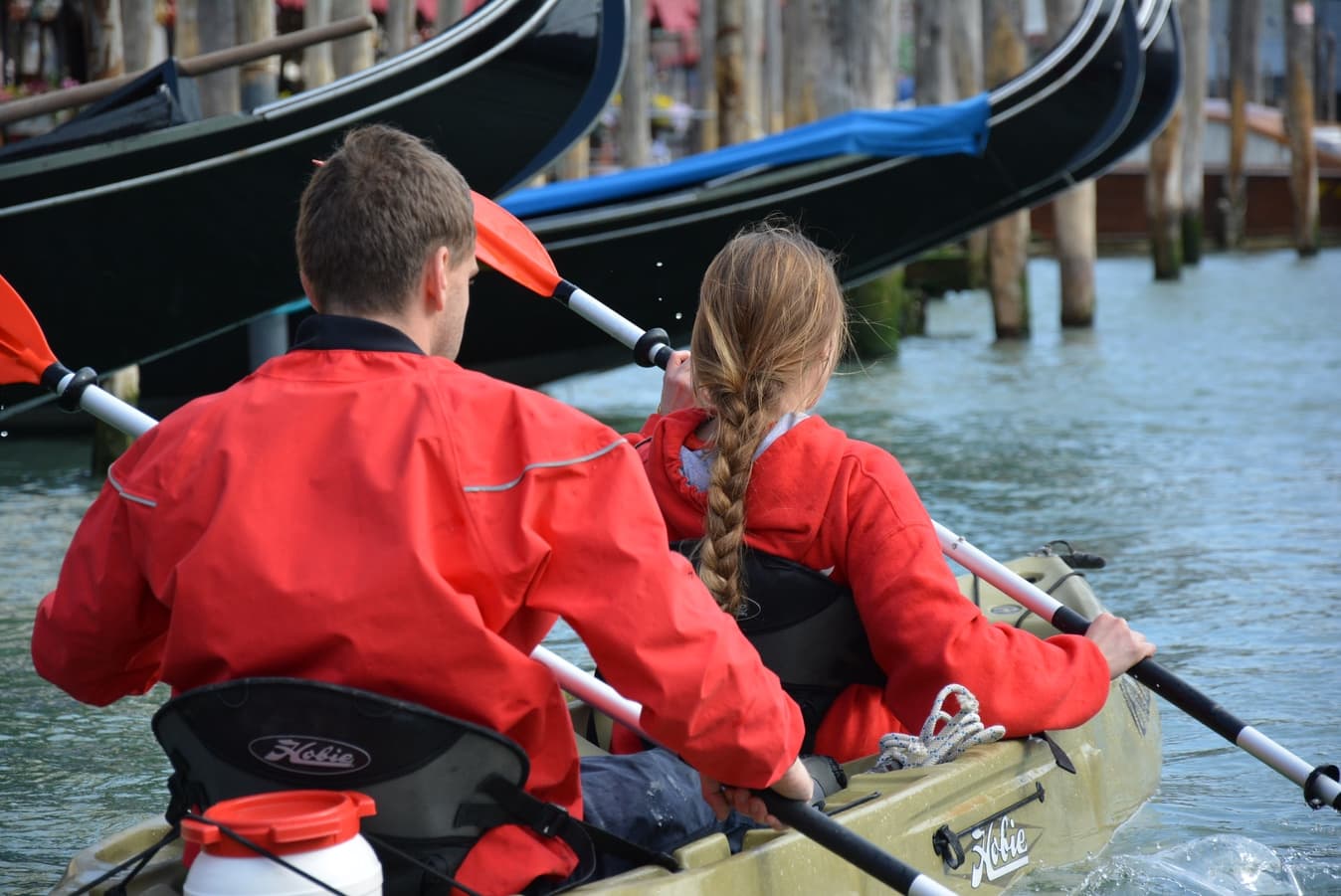 A couple paddling a tandem kayak past moored gondolas in Venice